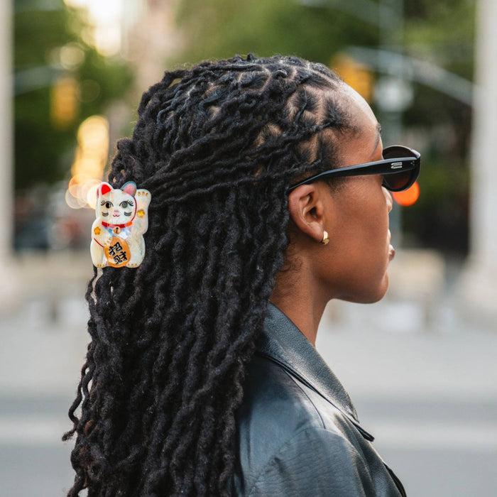 A woman in profile wearing black sunglasses and a black leather jacket, with a small white and orange Maneki-neko (lucky cat) hair clip holding her long black dreadlocks in place.