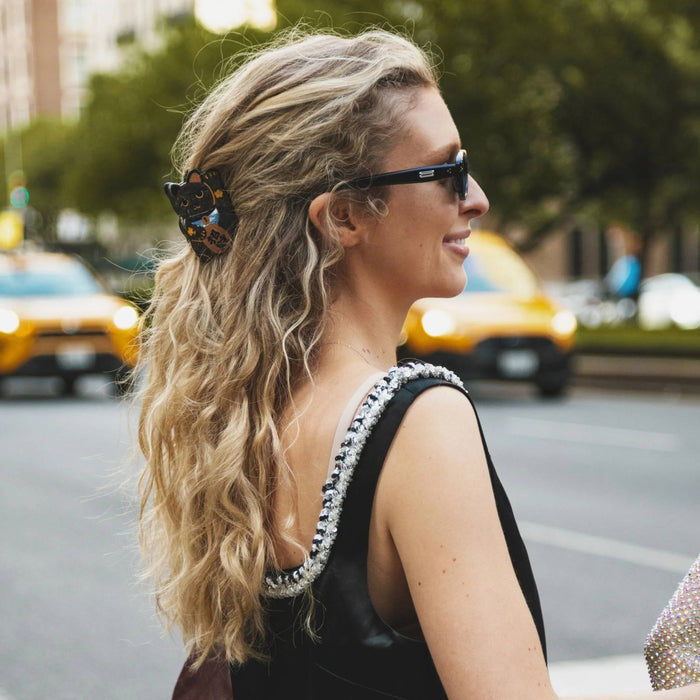 A woman in a black dress with a beaded strap and a black butterfly hair clip, photographed from behind over her shoulder in an outdoor city setting with yellow taxis in the background.