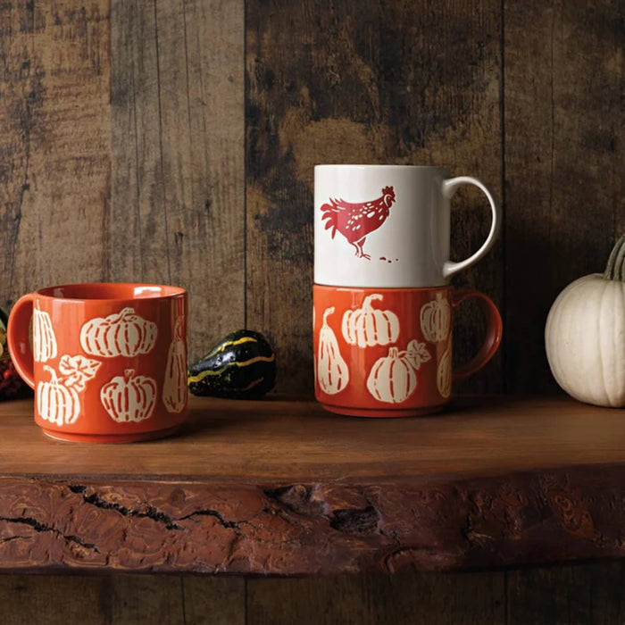 Two ceramic mugs with an autumn harvest theme stacked and displayed on a rustic wooden shelf with gourds, alongside a single mug to the left. The mug on the left and on the bottom of the stack is orange with wax relief gourd pattern and the mug on top of the stack is white with a red rooster.