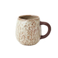 A stoneware mug with a textured, organic shape, featuring an embossed floral pattern, white reactive glaze and a contrasting dark brown handle, shown against a plain white background.