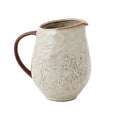 A handmade stoneware pitcher with a textured, off-white glaze featuring embossed botanical leaf patterns, a dark brown handle, and a brown rim, shown on a plain white background.