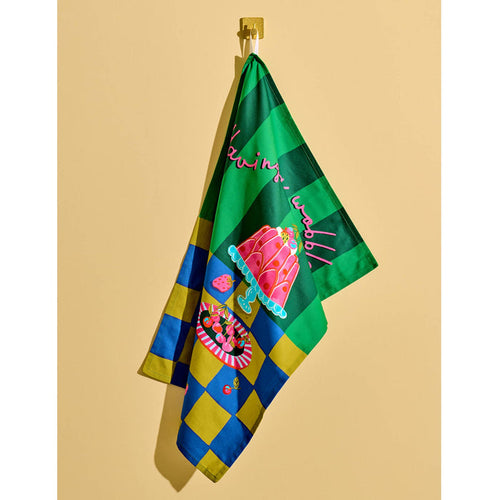 Tea towel with gelatin desserts on dessert plates on a blue and ochre surface with a two-tone green backdrop, hanging against a yellow background.
