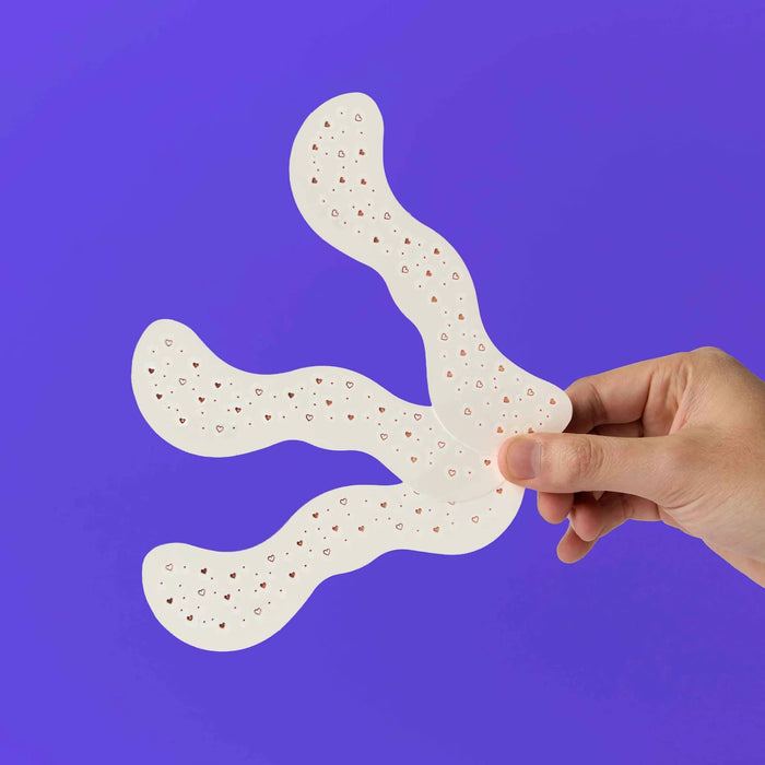 Hand holding 3 sheets of rose gold heart-shaped freckle tattoo sheets against a purple background.