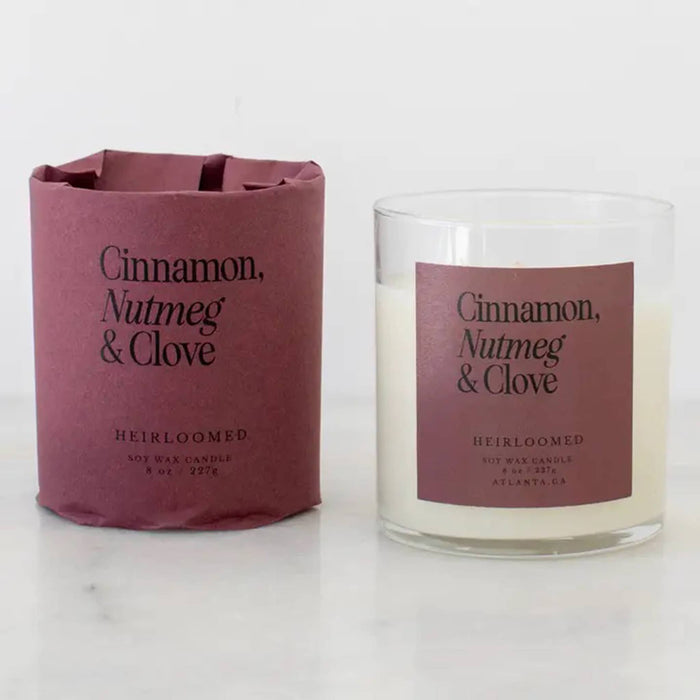A Heirloomed scented candle shown in a clear glass jar with cream-colored wax next to its matching dusty rose paper sleeve packaging. The label reads 'Cinnamon, Nutmeg & Clove'.