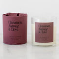 A Heirloomed scented candle shown in a clear glass jar with cream-colored wax next to its matching dusty rose paper sleeve packaging. The label reads 'Cinnamon, Nutmeg & Clove'.