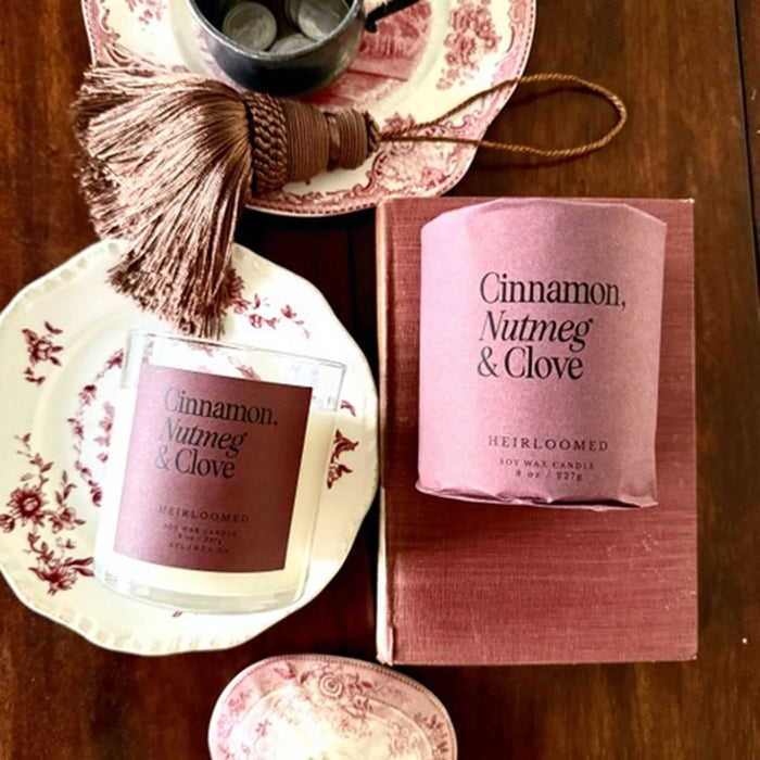 A candle product shown with its glass jar, pink paper sleeve, and matching pink box on a wooden table, styled with floral plates and a tassel.