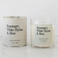 A soy wax candle in a clear glass jar next to its cream-colored paper wrapped packaging, both labeled 'Rosemary, White Thyme, & Moss' by Heirloomed, on a white marble surface against a white background.