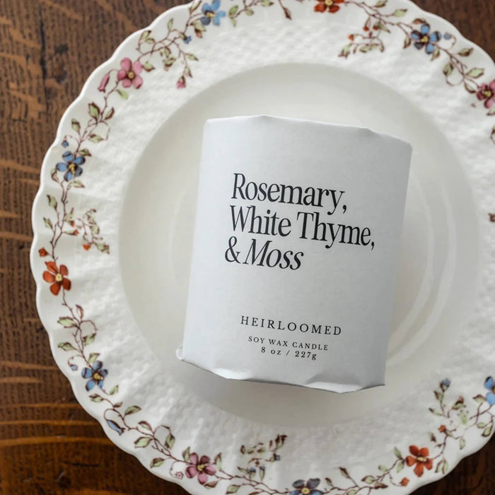 A white soy wax candle labeled 'Rosemary, White Thyme, & Moss' by Heirloomed, placed on a white ceramic plate with a floral border over a rustic wooden surface.
