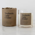 A soy wax candle in a clear glass jar next to its brown kraft paper packaging, both labeled 'Woodsmoke, Cedar & Ember' by Heirloomed, on a light marble surface against a white background.