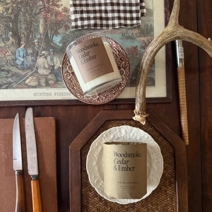 Two candles are styled on a rustic wooden table with knives, a checkered cloth, and antlers. The top candle is in a clear glass jar with brown label, and the bottom candle is in a white textured dish with a matching brown paper label reading 'Woods smoke, Cedar & Ember'.