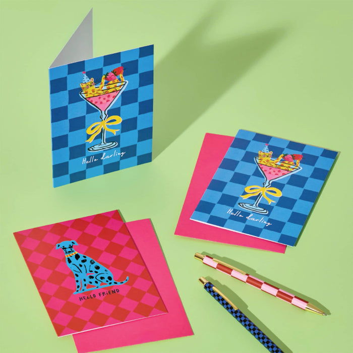 A set of colorful greeting cards arranged on a light green background, including a blue checkered card with a cat in a cosmo  illustration and the words 'Hello darling', a pink checkered card with a blue dog illustration and the words 'Hello friend', matching pink envelopes, and two decorative pens.