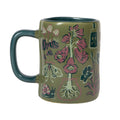 An olive green ceramic mug decorated with botanical and insect illustrations in pink, teal, and cream, shown on a plain white background.