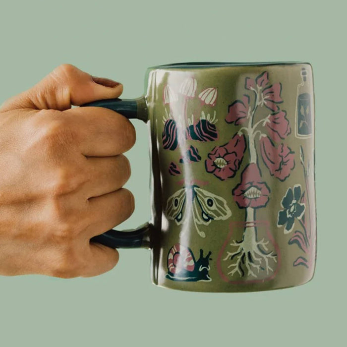 A hand holding a ceramic mug with a muted olive green base and a detailed botanical and insect pattern in pink, blue, cream, and dark accents, set against a solid sage green background.