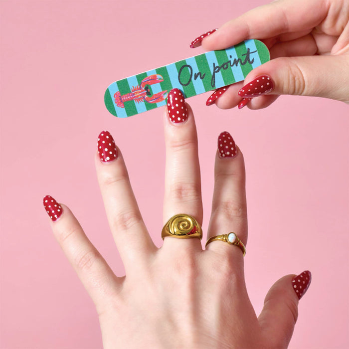 A close-up of a hand with red and white polka dot manicured nails holding a green and blue striped nail file with a lobster and 'On point' against a solid pink background.