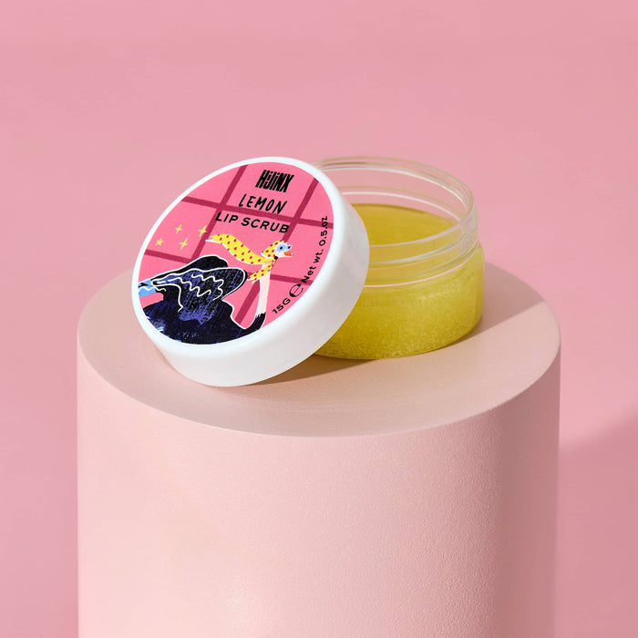 A clear jar of yellow lip scrub with its white lid leaning on top, set on a pink cylindrical pedestal against a pink background. The lid features a colorful graphic with the text 'HIJINX LEMON LIP SCRUB'.