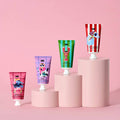 Four scented hand cream tubes displayed on pink cylindrical pedestals against a solid pink background. The tubes feature colorful, whimsical illustrations including an ostrich, lobster, cat, and dog.