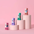 Four lip balm tubes in assorted colors and patterns are displayed on a pink step platform against a solid pink background. The tubes feature various designs including stripes, floral prints, and cartoon characters, with the brand name 'Hijinx' visible on the packaging.
