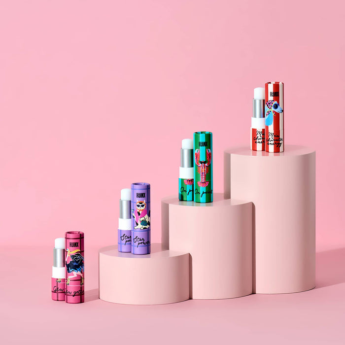 Four lip balm tubes in assorted colors and patterns are displayed on a pink step platform against a solid pink background. The tubes feature various designs including stripes, floral prints, and cartoon characters, with the brand name 'Hijinx' visible on the packaging.