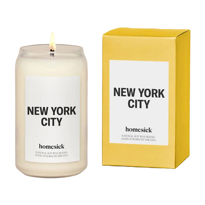 Candle in a clear glass can-shaped tumbler lit, white panel and "new york city" with "homesick" in black lettering beside yellow gift box with white panel and "new york city" and "homesick" with black lettering.
