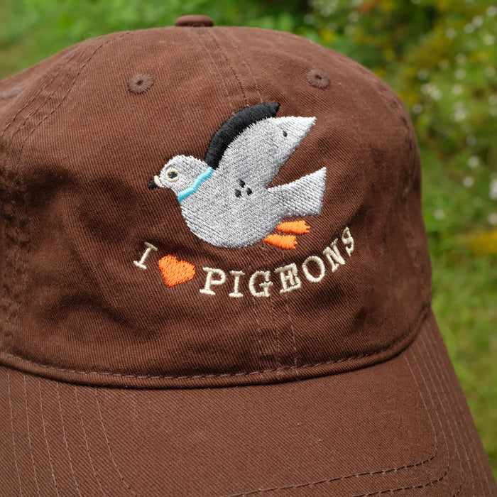 Brown cap with pigeon design and 'I ❤️ pigeons' text on a blurred grass background.