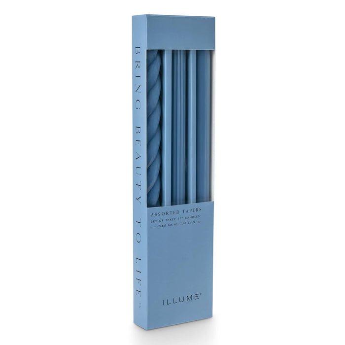 A tall rectangular box of ILLUME assorted taper candles in blue, shown on a clean white background. The box has a clear window revealing the tapers inside.