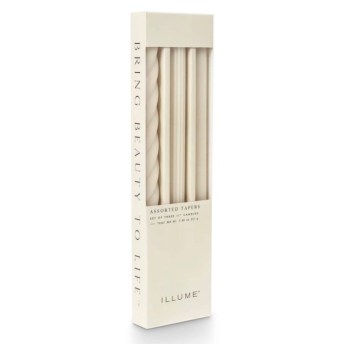 A tall rectangular box of ILLUME assorted taper candles in an ivory color, shown on a clean white background. The box has a clear window revealing the tapers inside.
