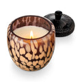 A lit candle in an acorn shaped amber glass vessel with beige speckles, shown with a black ribbed lid, on a white background.