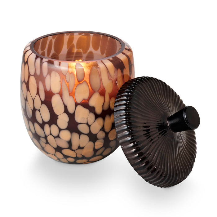 A decorative amber glass jar candle with a matching ribbed lid, shown on a clean white background. The jar features a mottled pattern of dark brown and warm beige, and a warm orange glow is visible from the lit candle inside.