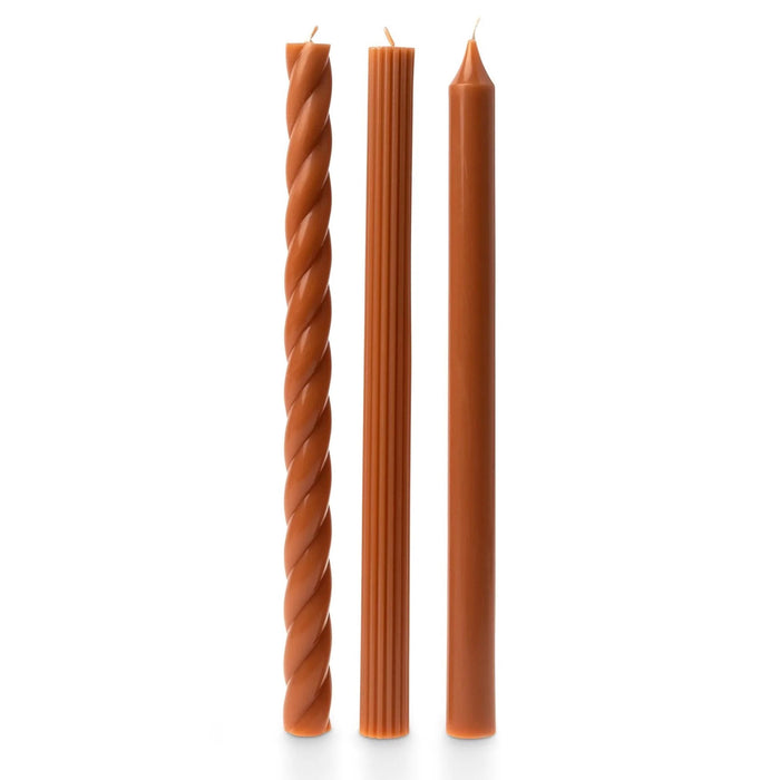 A set of 3 tall dark orange taper candles in different textures, a twisted spiral, a ribbed and a smooth, standing upright on a white background.