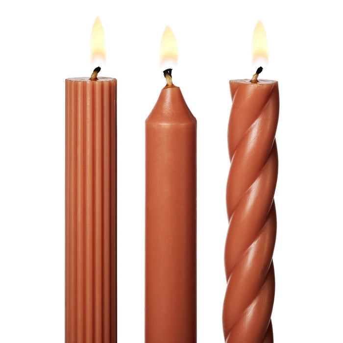 A set of 3 tall dark orange taper candles in different textures, a twisted spiral, a ribbed and a smooth, standing upright on a white background, detail of lit wicks.