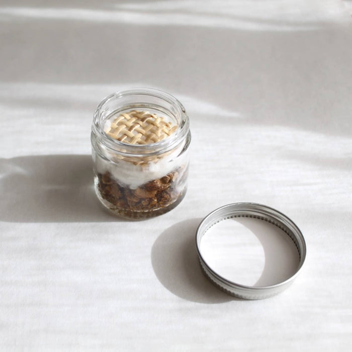Inspire and Create aromatherapy inhaler in a glass jar with woven top, lid beside it on a white surface.