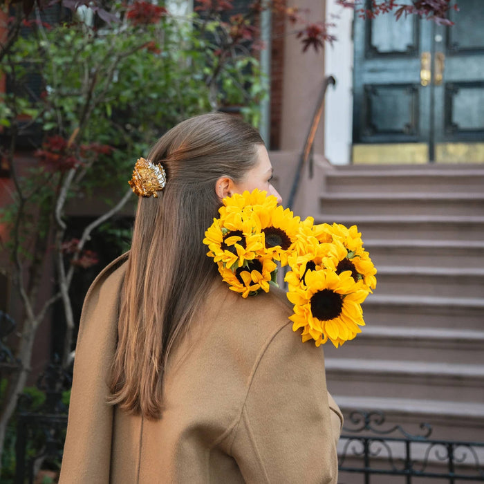 A woman in a tan coat is shown from the back outdoors, wearing a leaf hair claw, holding bright yellow sunflowers over her shoulder. Photographed in front of a building entrance with steps and greenery.