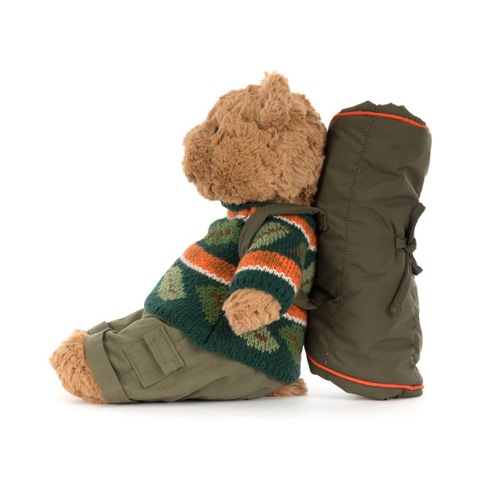 Jellycat Bartholomew Bear plush with a knit sweater in fall colors and a sleeping bag backpack, side view, on a white background.