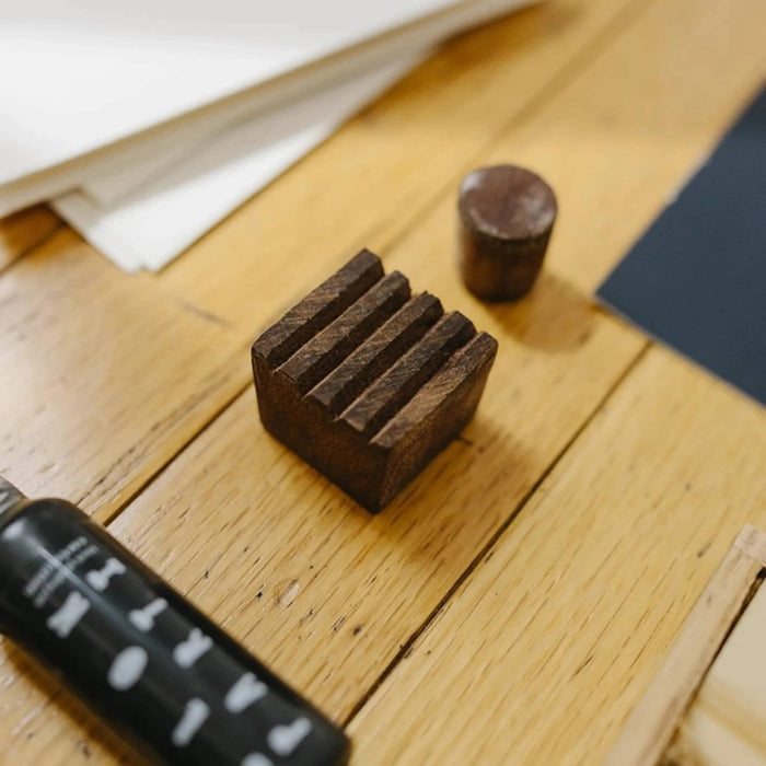 A dark brown wooden print block with a ridged top surface, shown on a light wood tabletop next to a small bottle of black ink.
