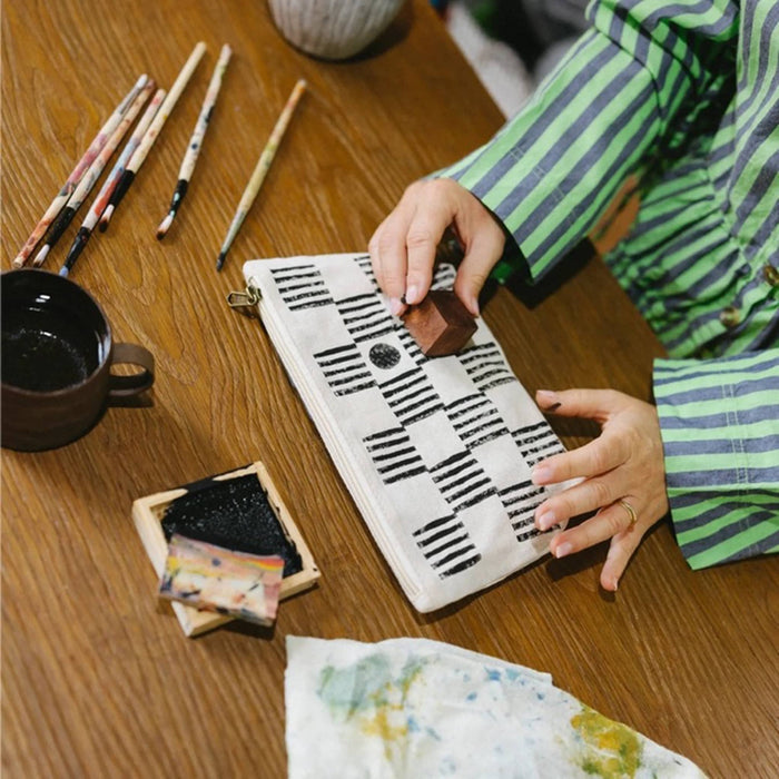 A person wearing a green and gray striped shirt is shown from the side, using a wooden stamp to print a black geometric pattern onto a cream canvas pouch. The scene is set on a wooden table with traditional block printing tools, including brushes, ink, and a cup.