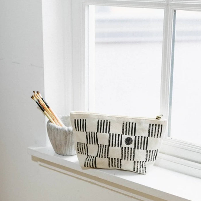 A canvas zip pouch with a black and white checkerboard pattern sits on a white windowsill next to a small ceramic cup holding pencils.