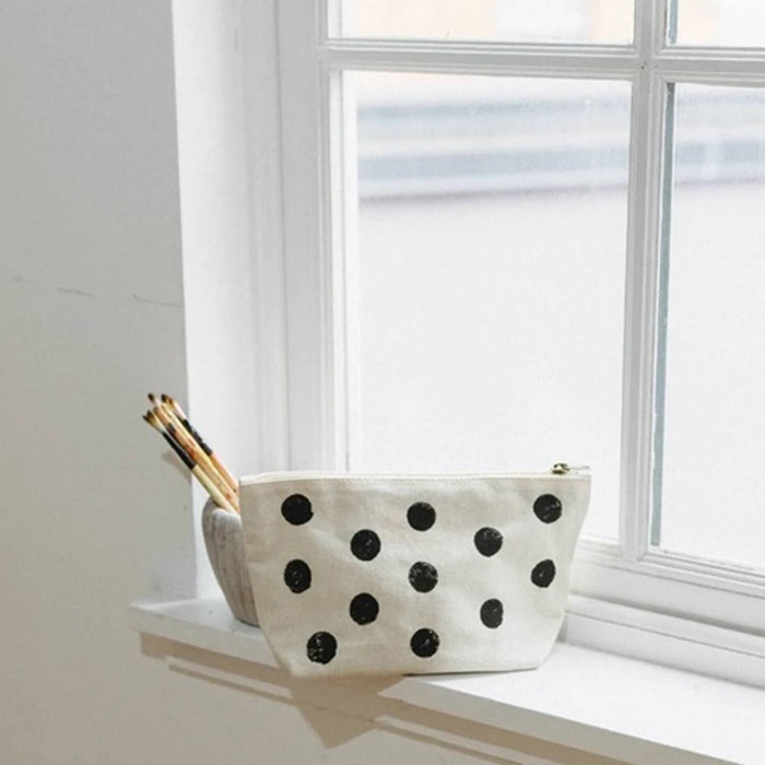 An off-white canvas zippered pouch with black polka dots sits on a white windowsill next to a small container holding paintbrushes.