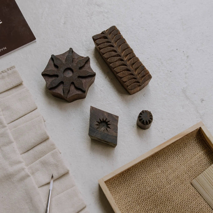 A styled flat-lay of carved wooden stamp blocks with floral and leaf motifs, arranged on a light textured surface alongside a canvas tote, a paintbrush, and a woven tray.