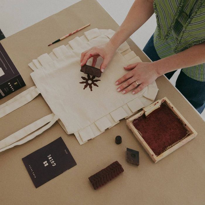 A person's hands are shown using a wooden block stamp to create a dark brown design on a cream canvas bag. The scene is set on a beige table with other printing tools, including a framed ink pad, ink blocks, and a printed card.