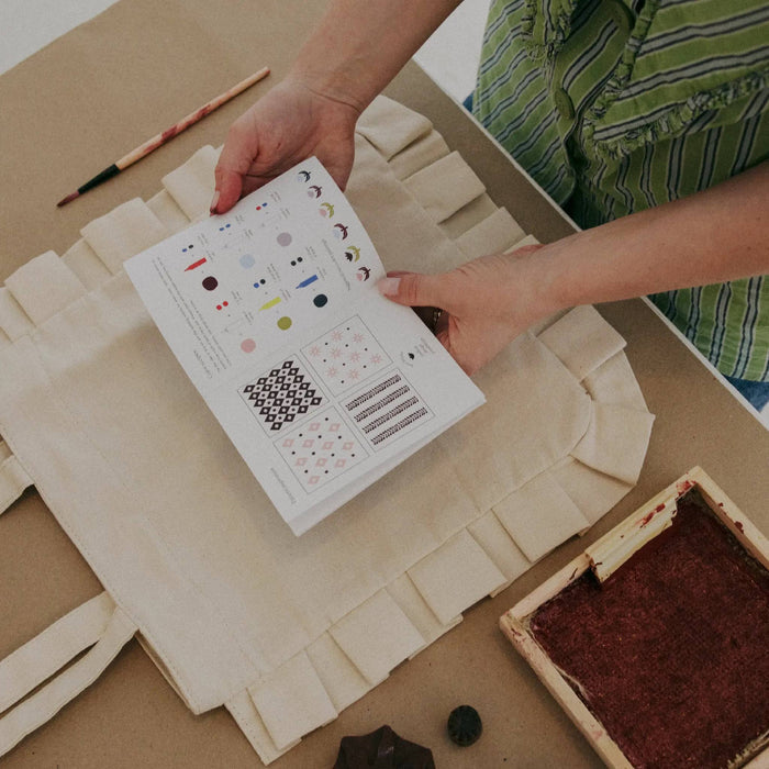 A person holds a printed guide book over a large cream canvas tote bag laid on a tan surface. The scene includes a green striped shirt, a paintbrush, and a red ink pad, suggesting a craft or apparel design process.