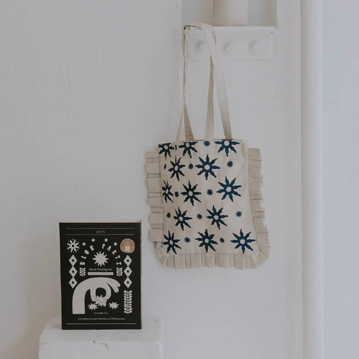 Ruffle tote bag with a blue block print pattern, hanging on a white wall next to block print kit packaging.
