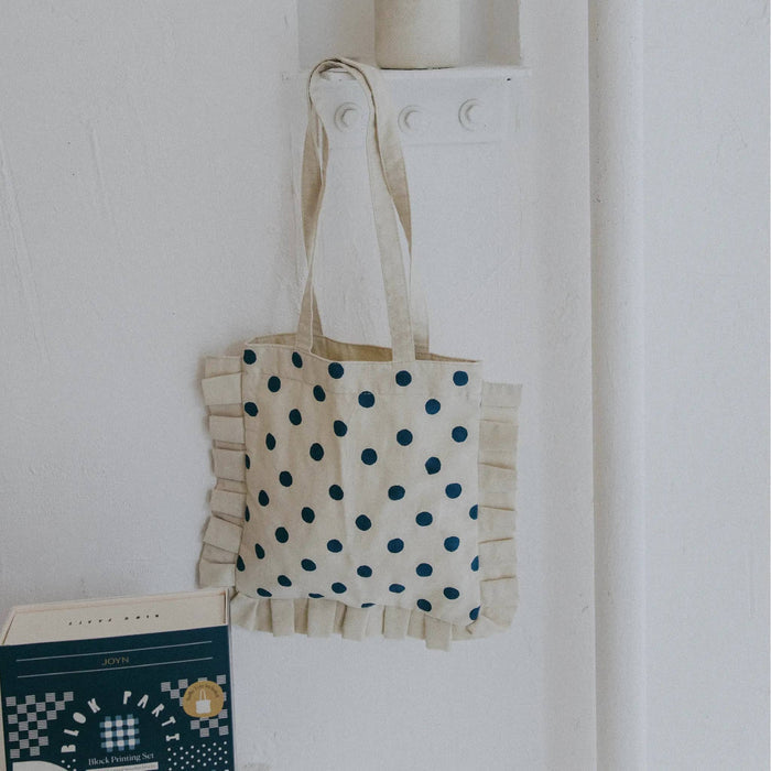 A cream canvas tote bag with dark blue polka dots and ruffled trim, hanging on a white wall hook.