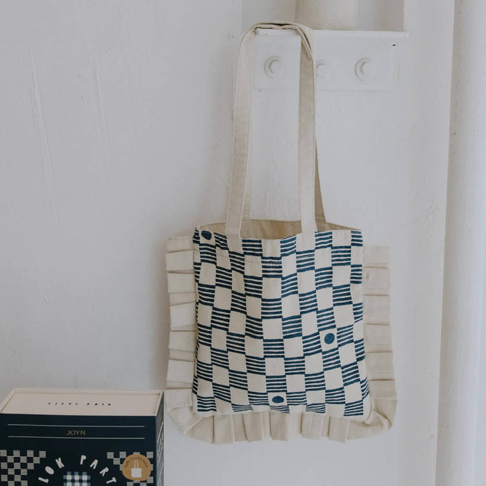 Cream ruffle tote bag with checkered pattern hanging on a white wall.