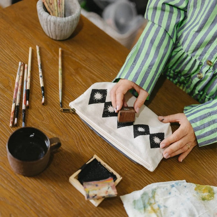 A person wearing a green and gray striped shirt sits at a wooden table, using a wooden block to print a black and white diamond and star pattern onto a canvas pouch. The scene is styled with traditional block printing tools, including brushes, and an ink tray.
