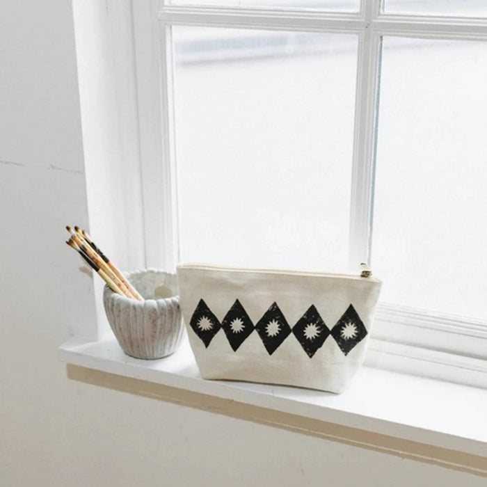 A cream canvas zip pouch with a black diamond pattern sits on a white windowsill next to a small textured cup holding pencils.
