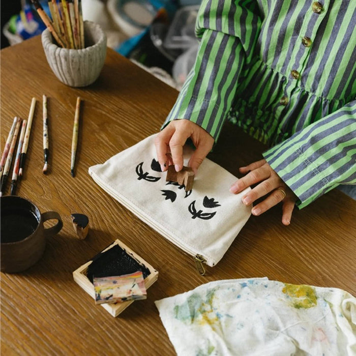 A person wearing a green and gray striped shirt is shown from the chest down, placing a small wooden block onto a cream canvas bag with black floral designs. The scene is set on a wooden desk surrounded by art supplies, including brushes, ink, and a cup of tea.