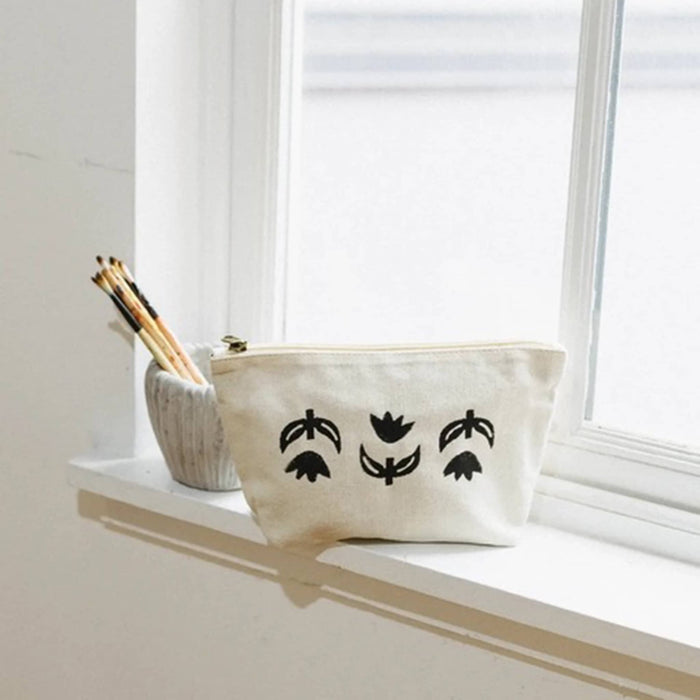 An off-white canvas pouch with three black stylized leaf and flower motifs sits on a white windowsill next to a textured ceramic pencil holder with pencils.