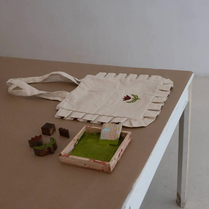 A cream canvas tote bag with ruffled edges, featuring a small block printed red flower with green leaves, displayed on a brown table next to small wooden toy blocks and a miniature ink tray filled with green ink.