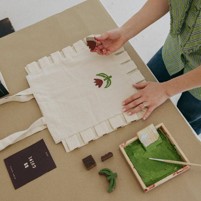 A person's hands are shown stamping a green and red tulip flower onto an off-white canvas ruffled tote bag. The scene is set on a light brown table with a small ink tray filled with green ink, a printing block, and a booklet on a beige surface.