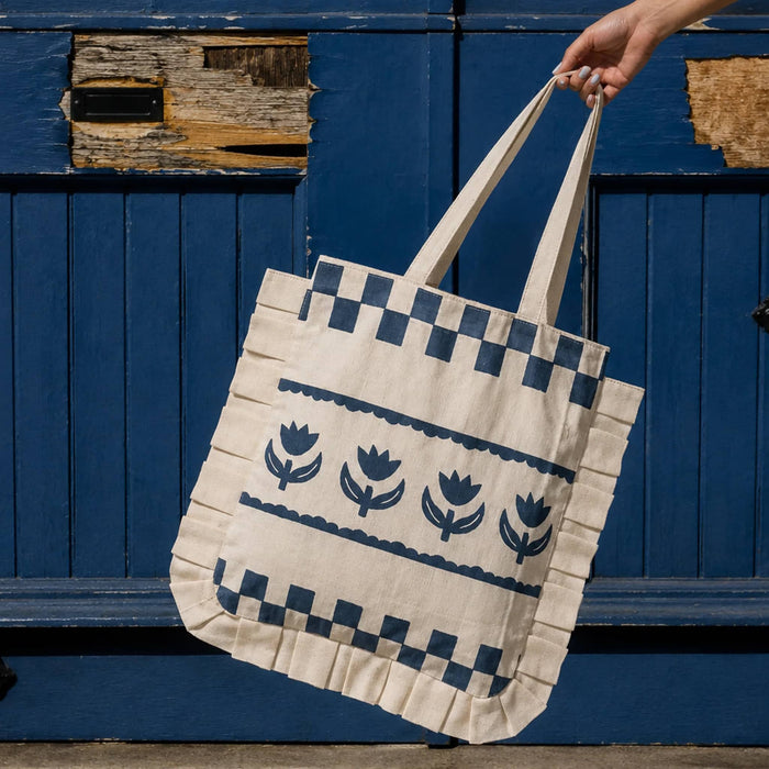 A cream canvas ruffled tote bag with dark blue printed tulip patterns, held by a hand in front of a rustic blue wooden door.